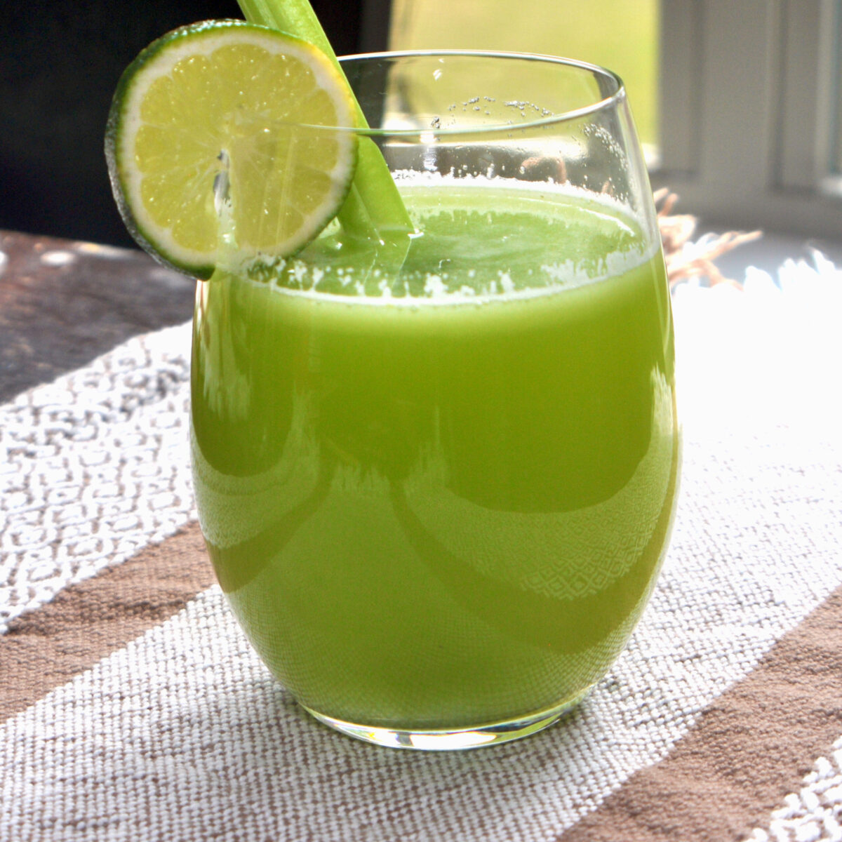 celery juice in a stemless glass with a celery stalk in the glass and a slice of lime on the rim.