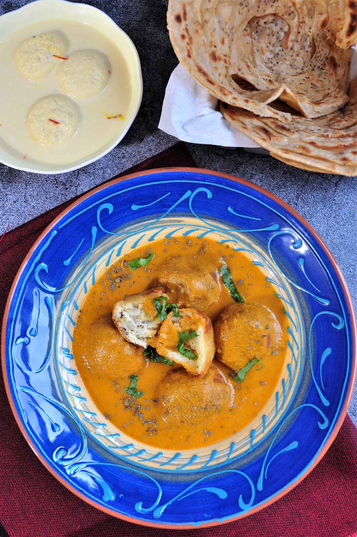 malai kofta served in a bowl with paratha and rasmalai in background.