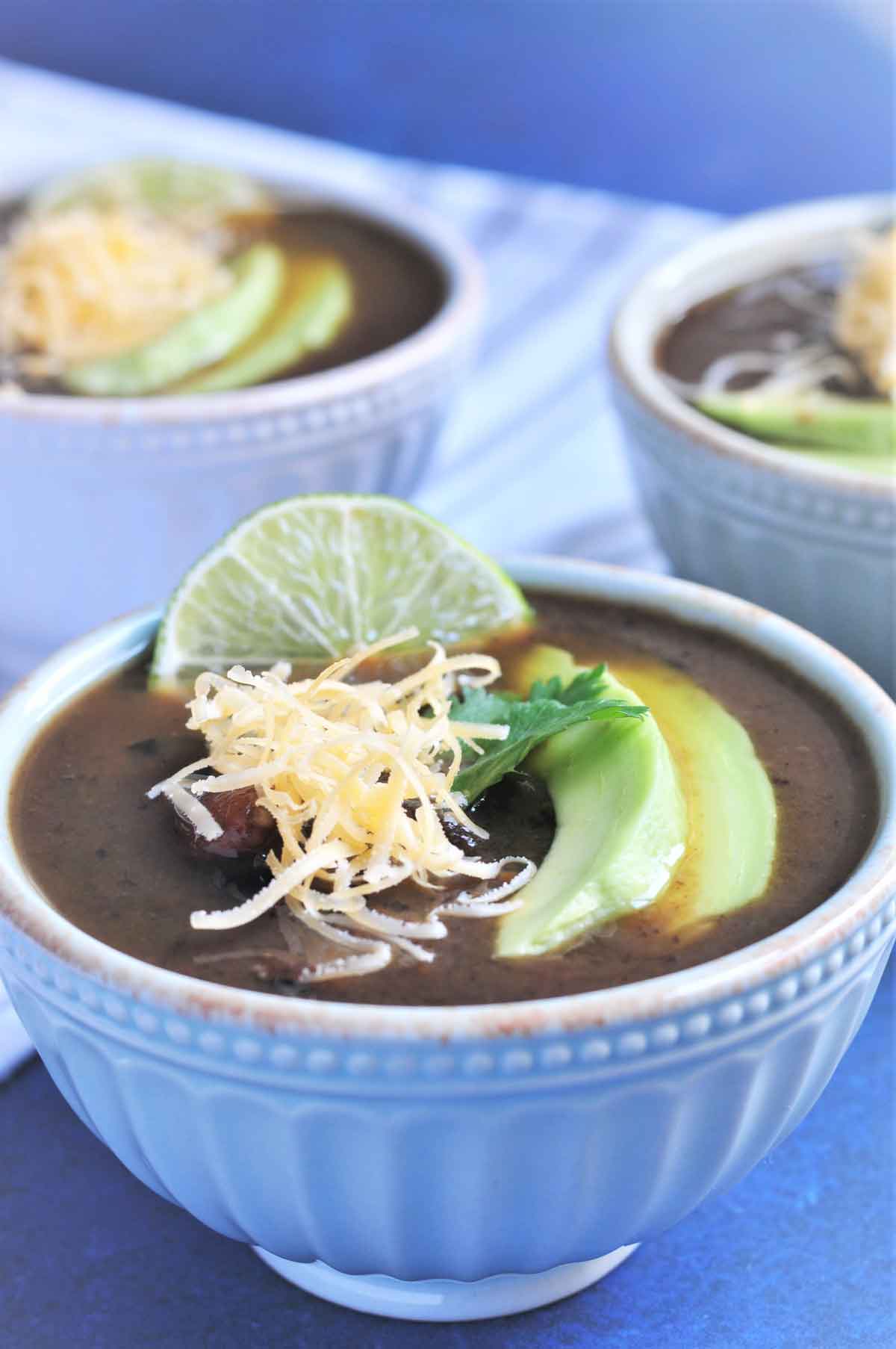 Black Beans Soup in a bowl and garnished with lemon, avacado slice and shredded cheddar cheese.